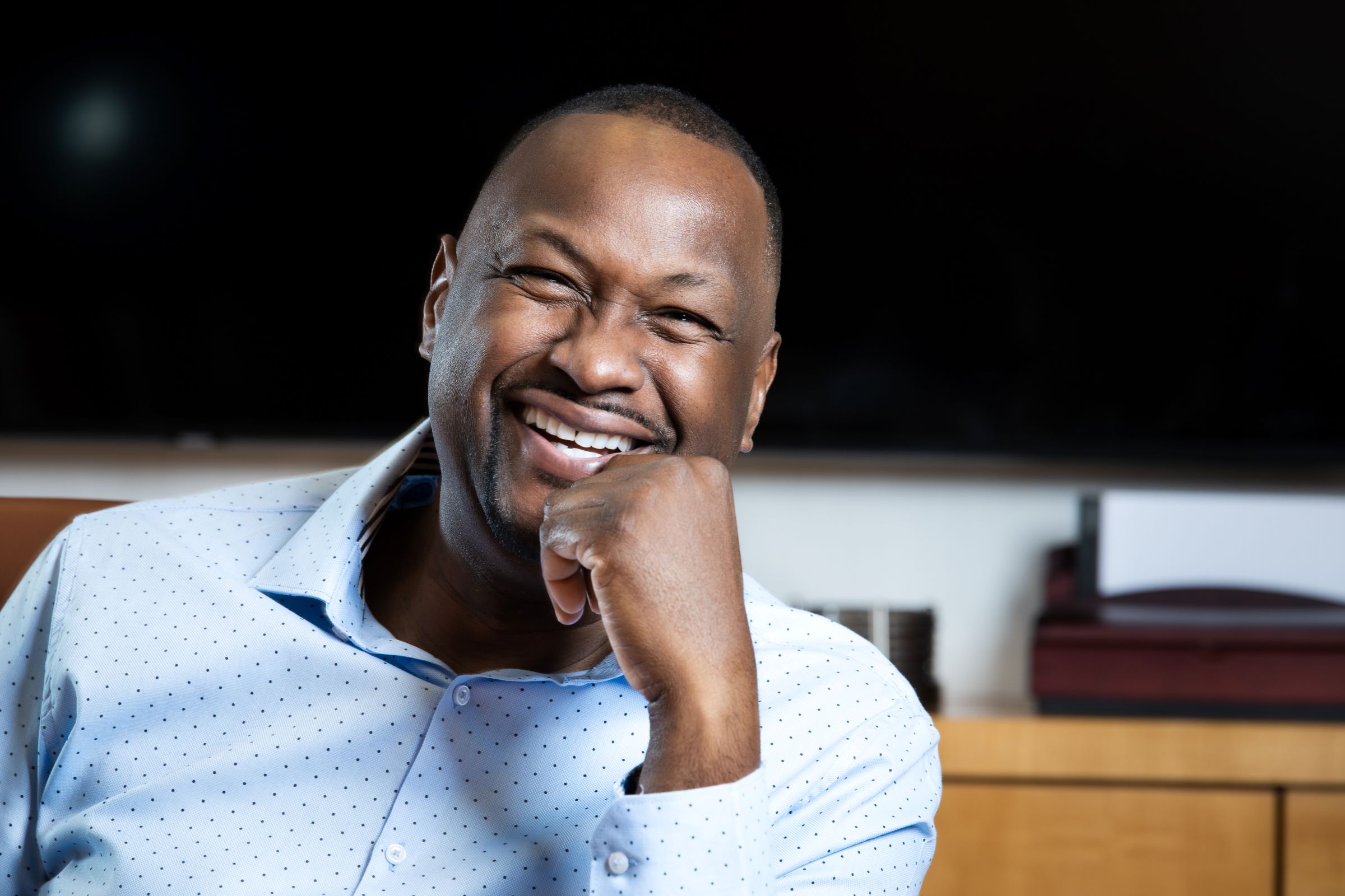 Picture of Dr. Kevin John smiling and sitting at desk in conference room.