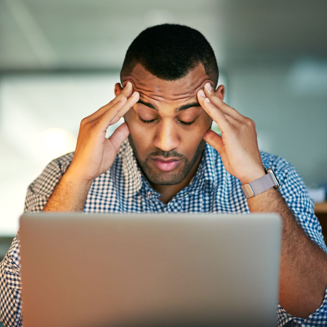 Stressed leader sitting at desk in front of laptop with head down and hands rubbing his temple.